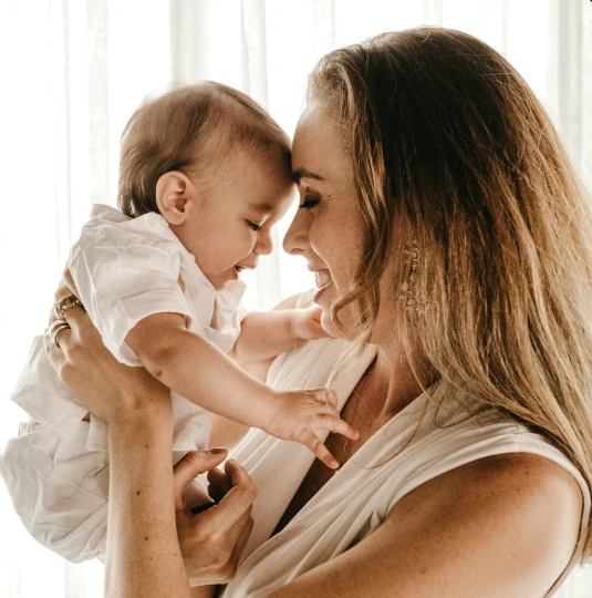 Mother lifting her baby up close, forehead to forehead, both smiling in bright window light